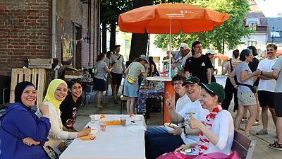 sechs Personen sitzen an Tisch draußen, lachen, weiß und People of Colour, im Hingergrund mehrere Menschen