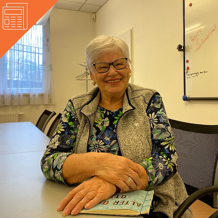 Das Foto zeigt eine ältere Frau mit kurzen weißen Haaren und einer Brille. Sie sitzt lächelnd an einem Tisch und hat die Arme locker übereinandergelegt. Die Frau trägt ein buntes, blumengemustertes Oberteil und eine graue Weste darüber. Auf dem Tisch vor ihr liegt ein Fotoalbum mit dem Titel „ALTER ORT“. Im Hintergrund ist ein heller Raum zu sehen: ein Fenster mit Vorhang, eine Garderobe und ein Whiteboard. Die Atmosphäre wirkt freundlich und einladend.