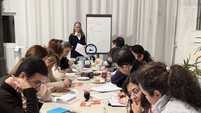 Menschen sitzen um eine lange Tafel und sind in ihre Lernunterlagen vertieft. Am Ende der Tafel steht die Vortragende neben einem Flipchart.