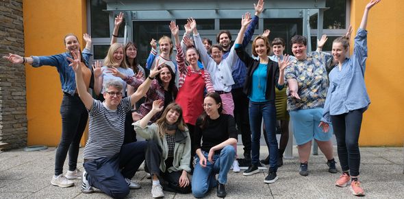 Die Mitarbeiter*innen des GB*Teams stehen vor dem Lokal am Max-Winter-Platz und strecken die Hände in die Höhe