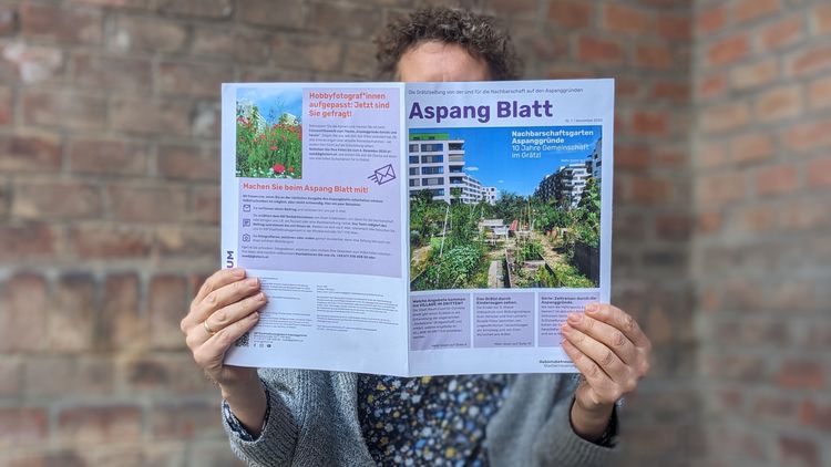 Frau sitzt vor Backsteinfassade auf einem türkisen Holzklappstuhl und liest die erste Ausgabe des Aspang Blatts -- die Nachbarschaftszeitung verdeckt ihr Gesicht.