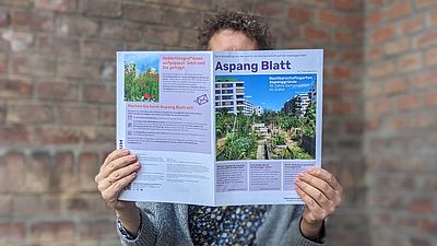 Frau sitzt vor Backsteinfassade auf einem türkisen Holzklappstuhl und liest die erste Ausgabe des Aspang Blatts -- die Nachbarschaftszeitung verdeckt ihr Gesicht.
