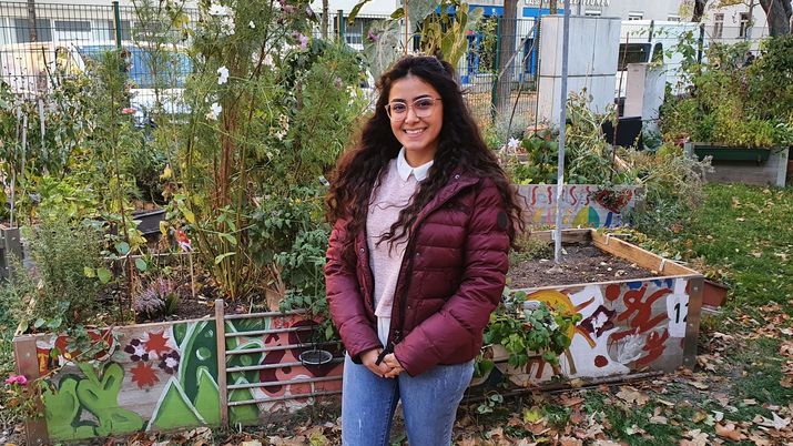 Junge Frau mit Brille, langen dunklen Haaren und bordeauxfarbener Jacke  steht in einem Garten und lächelt in die Kamera