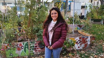 Junge Frau mit Brille, langen dunklen Haaren und bordeauxfarbener Jacke  steht in einem Garten und lächelt in die Kamera
