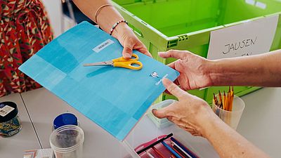 Ein Schulheft und eine Schere werden von einer Person an eine andere übergeben.