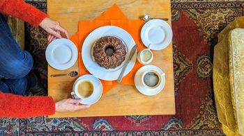 Ein gedeckter Kaffeetisch aus Holz. Ein Stracchiatellaguglhupf steht auf einem weißen Teller auf orangenen Servietten. Auch zwei Kaffeetassen sind angerichtet. Zwei Hände bewegen eine der Kaffeetassen und Untersetzer.