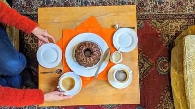 Ein gedeckter Kaffeetisch aus Holz. Ein Stracchiatellaguglhupf steht auf einem weißen Teller auf orangenen Servietten. Auch zwei Kaffeetassen sind angerichtet. Zwei Hände bewegen eine der Kaffeetassen und Untersetzer.