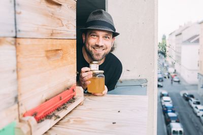 Mann mit Hut und einen Glas Honig grinst hinter einem Bienenstock hervor