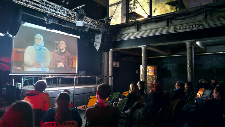 Ein Publikum schaut auf eine Leinwand, auf der ein Dokumentarfilm läuft.