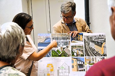 Ein Mann halt eine Tafel mit Bilder und einem Entwurfsplan, mehrere Personen sind im Bild zu sehen, die informiert werden.